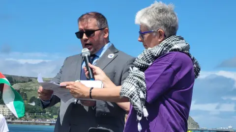Steve Witherden is speaking into a microphone held up by a protester wearing a keffiyeh-style scarf around her neck. A Palestinian flag can be seen to the left of the picture, with Llandudno's skyline in the distance with blue skies and clouds.