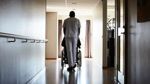 Getty Images Corridor of a nursing home, with bare walls to the right and left. A person in grey scrubs is wheeling a person in a wheelchair away from the camera. They are heading towards brown curtains on either side of the corridor.