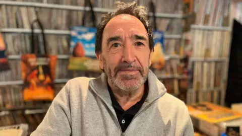 A man with dark hair and a beard in a grey zipped top with a black T-Shirt underneath stands in front of a huge array of vinyl records
