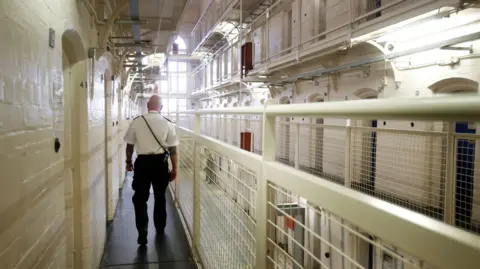 An image from inside HMP Barlinnie in Glasgow, with a bald uniformed prison guard walking away from the camera along a narrow walkway, enclosed by white metal railings.