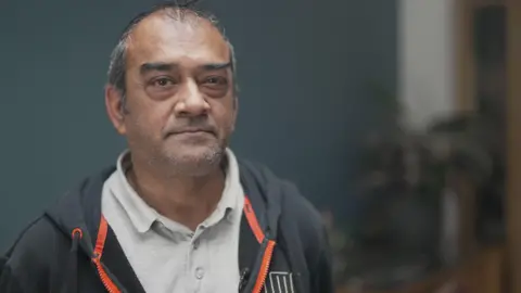 A man wearing a grey polo shirt and a blue hoodie sits looks at the camera