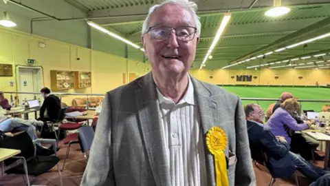 Roger Evans is wearing a white polo shirt with a grey jacket on top. He is standing in the hall used for the election count