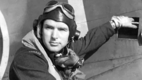 A black and white photo of Lieutenant John Saville in front of a plane. He is wearing his uniform which includes a pilot's cap and goggles. 