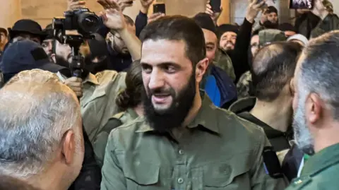 Reuters Ahmed al-Sharaa, previously known as Abu Mohammed al-Jolani, the leader of HTS, speaking to a group of supports inside the Ummayad Mosque in Damascus. Many people are taking photographs of him. He is wearing a green military-style shirt.