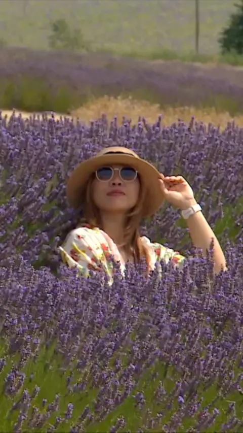 A woman holds her hand to her hat as she sits in a field of lavender.