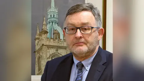 Presbyterian Church in Ireland Rev Dr David Allen pictured infront of a painting of a building. He has short grey hair and a beard, wears dark framed glasses, a navy suit and blue shirt with a black and blue paisley tie.