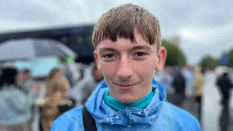 A boy looking at the camera. He has dark hair and dark eyes. He is wearing a light blue jacket with a turquoise jumper underneath. The background is blurred and the sky is grey.