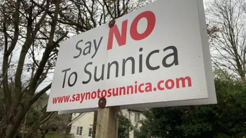 Shaun Whitmore/BBC A sign saying Say No to Sunnica in back and red letters on a white background in front of winter trees and a white house