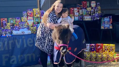 A little boy is sat on a pony and smiling. A woman is standing next to him and hugging him. There are piles of easter eggs behind them
