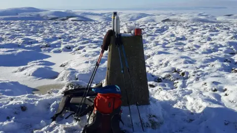 Patrick Norris A trig point sits in a snowy landscape which stretches in all directions. On the top of the trig point is flasks and a cup and backpacks are placed at its base 