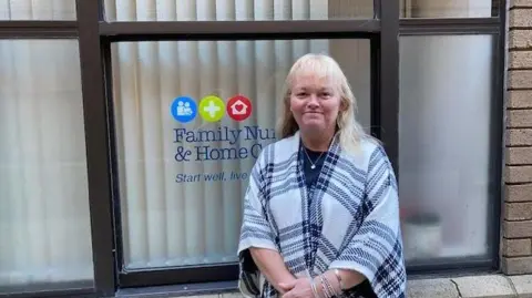 Mandy Le Tensorer stands in front of the Family Nursing and Home Care office window. A logo for the charity is visible behind her. She has shoulder length blonde hair and is wearing a plaid shawl.