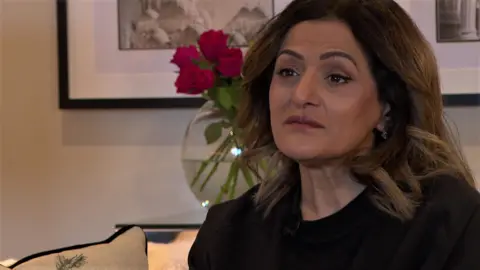 A woman with dark hair and brown eyes sits on a sofa. She is wearing a black top and jacket and earrings. Behind her are two artworks on a wall and a vase of pink roses.