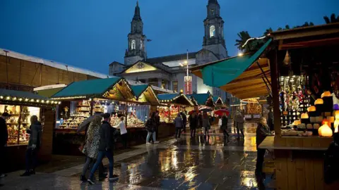 Getty Images Leeds Christmas market at night, in the rain, showing people walking past wooden cabins selling Christmas-themed gifts with Leeds Civic Hall in the background