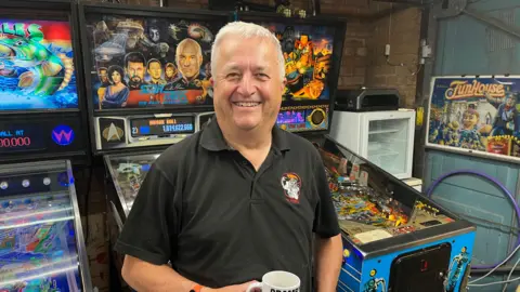 Mark Squires,  with short white hair, wearing a grey polo shirt, standing in front of three colourful pinball tables