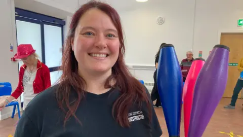 Elly Cribbens is standing in the exercise room wearing a black shirt. She has red hair and is smiling at the camera.