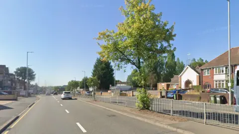 A street view image of a dual carriageway. It is a sunny day, with a few cars on the road, and a tree standing at the barrier in the centre of the carriageway.