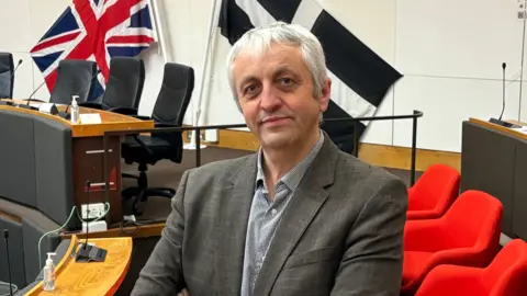 BBC Dick Cole has grey grey hair and wears a grey jacket and black and white checked shirt. He stands inside the Cornwall Council chamber with seats and the Union and Cornish flags behind him. 