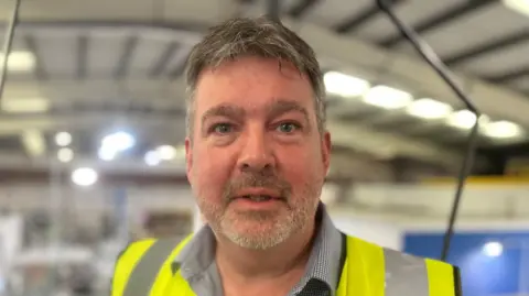 A middle-aged man wearing a hi-vis jacket looking at the camera - he has a grey bear and greying short hair and blue eyes. You can see in the background he is in a factory setting.