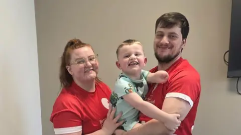 Libby and George stand in front of a white wall wearing their London Marathon T-shirts and holding son Ritchie between them. 