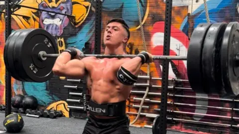 Joel McManus A topless man holding a weightlifting bar with several heavy weights at shoulder level