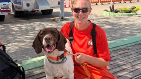 Lancashire Fire and Rescue Service A female firefighter, with blonde hair scraped back and sunglasses, smiles as she poses with her brown and white spaniel sniffer dog on a bench in Venezuela.