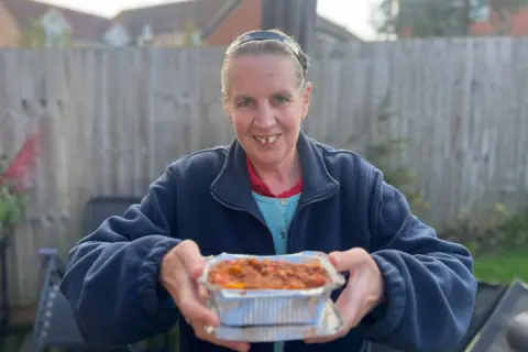 A woman wearing a blue zip-up jacket is holding a silver tray filled with spaghetti bolognaise. She is looking directly into the camera smiling. She is standing in the garden with a grey fence behind her. 