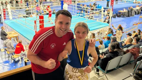 Isabel Mitchell Two people are posing in a boxing stance in front of a boxing ring at a competition. The woman is wearing a blue boxing vest and white shorts with the name Izzy on the front. She has a gold medal aroud her neck and is smiling. Her coach is standing next to her wearing a red T-shirt. He has his right fist clenched like a boxer and is smiling. 