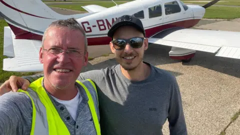 Family handout An older man (left) and a younger man wearing a cap and shades in front of a light aircraft on a runway