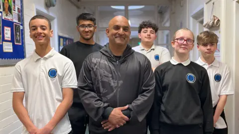 Aimran Majid stands in a classroom corridor surrounded by the students he teaches at Lindsworth School. He is wearing a black jacket and trousers, the five male and female students are wearing their school uniform. 