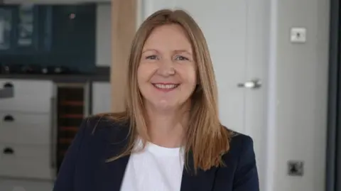 Shaun Whitmore/BBC Louise Brown smiles at the camera inside a rental holiday home. She has long red hair and wears a navy suit jacket with a white T-shirt underneath. 