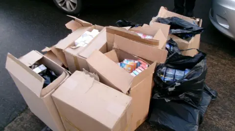 Multiple cardboard boxes and a few binbags on a road. They are full of fake illegal tobacco products.