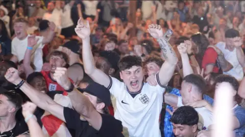 An England fan, in a white shirt, is held aloft by friends as he screams in delight