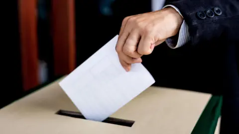 A ballot sheet being put in a box. The person has a black suit and white shirt.