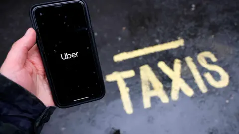 A person holding an apple i-phone with the uber app open, standing next to a yellow sign saying "taxi" on the road.