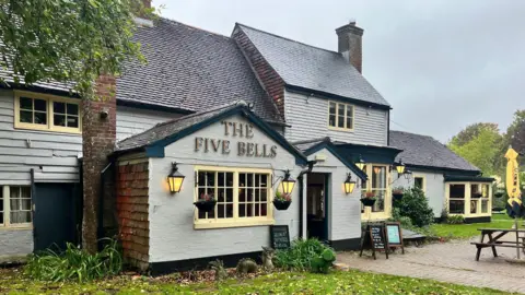 The Five Bells A pub building. It has grey outer walls and a black room. There is a large sign on the outside which says 'THE FIVE BELLS'. 