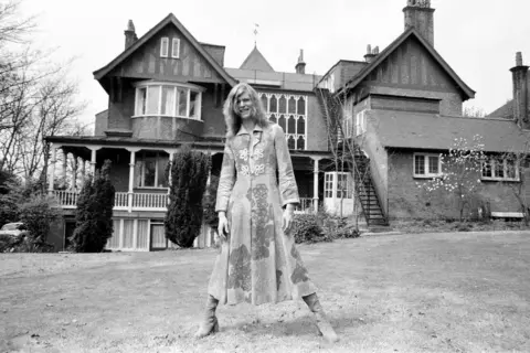 Mirrorpix via Getty Images A black and white photo David Bowie with long, fair hair wearing a long dress coat and suede boots while standing on a lawn in front of a sprawling Victorian villa featuring a round bay window, verandas and balconies.