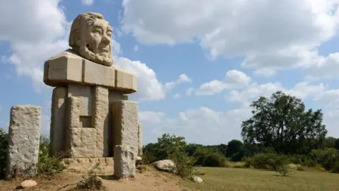 Corbis via Getty Images A stone bust of Paul Kruger sits atop some concrete slabs. It is in the Kruger National Park and greenery and trees can be seen around it.