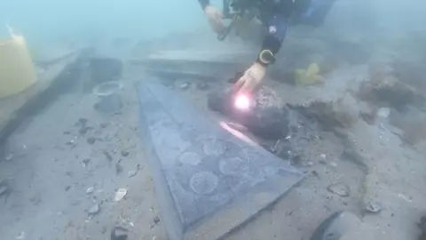 Bournemouth University A diver underwater shining a torch on the exposed grave slab seen above, which lies partially buried in the sand, surrounded by excavating equipment like small buckets. 