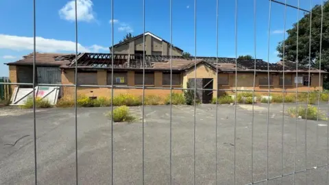 Local Democracy Reporting Service A boarded-up building with a hole in the roof and overgrown weeds outside in the car park pictured from behind a security fence.