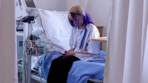A young woman with blonde and purple coloured hair sits looking at her phone on a hospital bed. She wears a hospital gown and monitors can be seen in the room. 