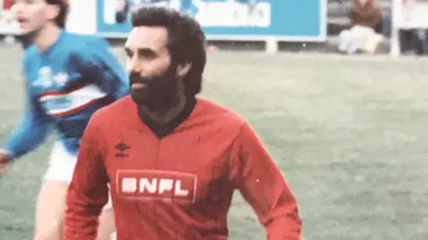 Workington AFC George Best in a Workington Reds shirt playing at Borough Park. He has black hair and beard and is caught in action, with another player in blue behind him running.