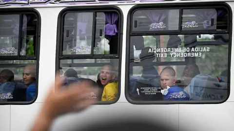 Federico PARRA / AFP via Getty Images Inmates look through the window of a bus as they are transferred from the Internado Judicial Barinas (Injuba) prison, in Barinas, Venezuela, on May 25, 2026. Their heads are shaven and they are wearing prison T-shirts, some of them blue, others yellow. One inmate appears to be shouting something to relatives gathered outside the prison.