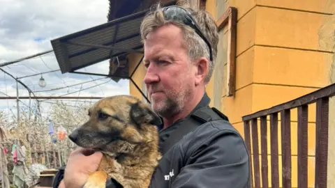 Paul Farthing is cradling a dog in his arms in front of an orange building. He has light brown hair and stubble, and is wearing a black jumper with sunglasses on his head.