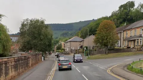 Google St Mary Street, Risca, with a residential area to the right, cars passing each other on the road, trees visible in front of the houses and forest in the background. 