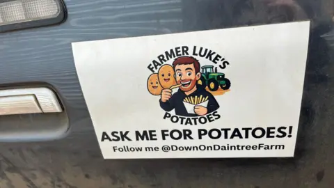 John Devine/BBC A magnetic sign on the side of a 4x4 vehicle that reads "Farmer Luke's Potatoes". It shows a cartoon image of Luke eating a bag of chips.