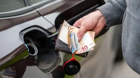A man holding euro notes and a wallet in front of an open petrol cap of a dark coloured car. 