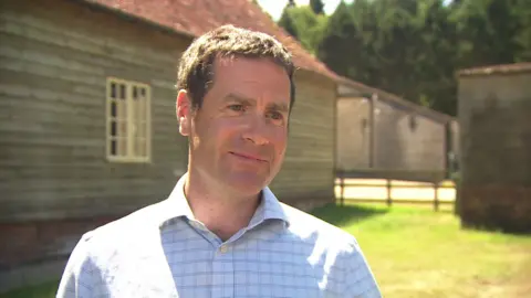 BBC A white man with brown hair standing outside a house in the rural countryside smiling.
