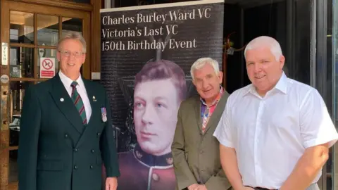 Three men stand next to a large sign advertising an exhibition. The sign has an old photograph of a man in soldier's uniform and reads: Charles Burley Ward VC Victoria's Last VC 150th Birthday Event. 