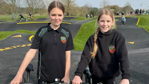 Gavin Kermack / BBC Two 12-year-old girls wearing black school uniformts stand with their bikes in front of the pump track.