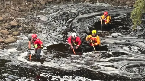 Rescue teams in hard hats and dry suits search the river Tyne in the Wylam area. The water is dark with foam swirling on the surface.